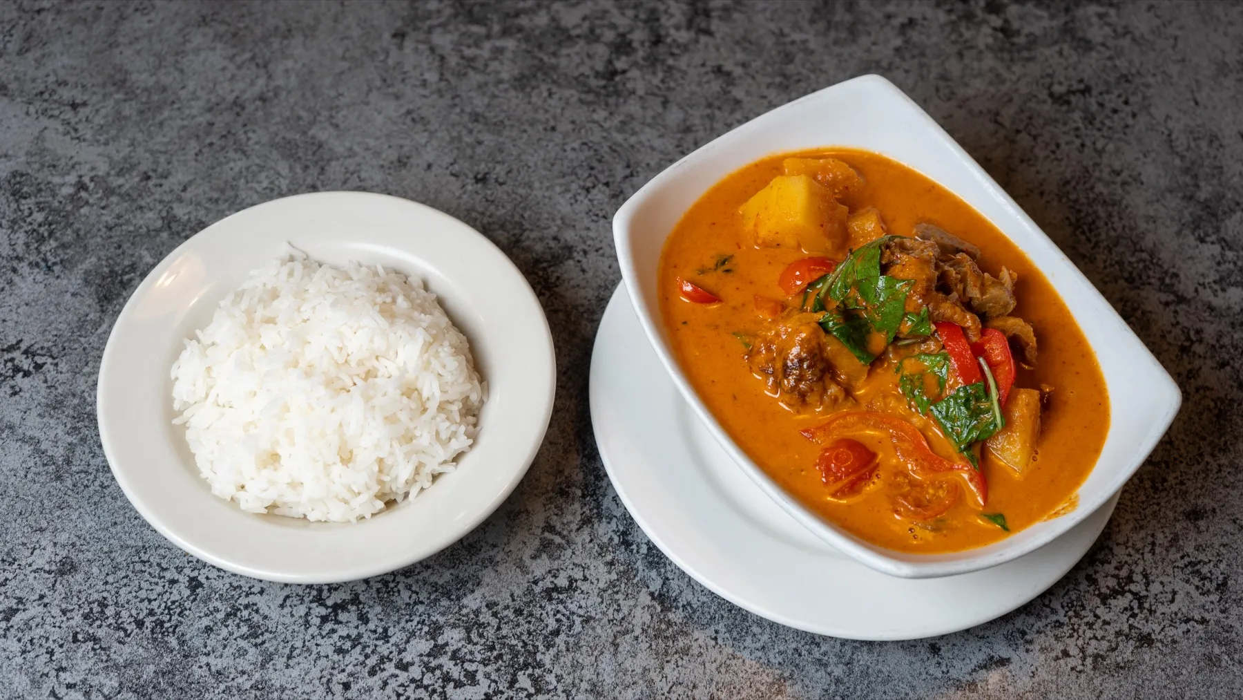 A bowl of orange Thai curry with vegetables and basil served beside steamed rice